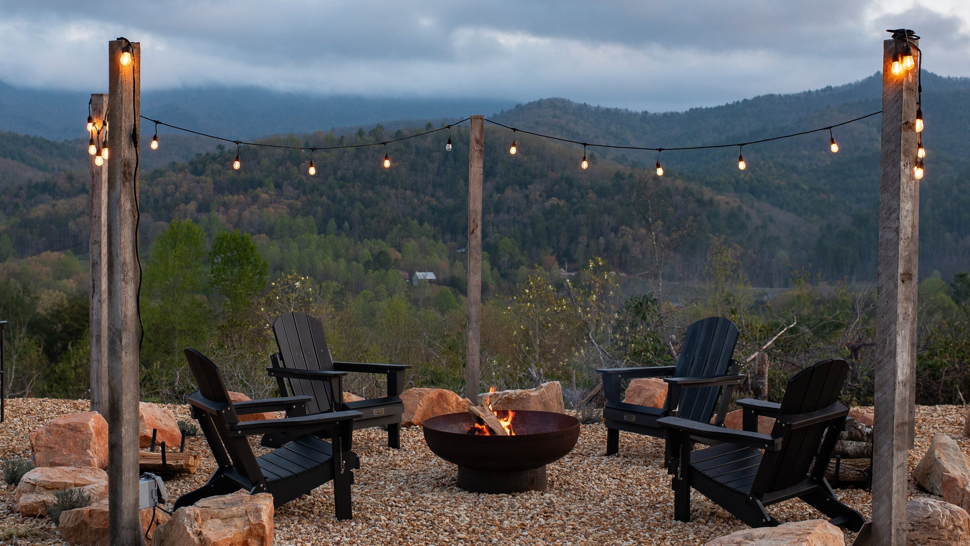 Outdoor setting with a fire pit and chairs under string lights with a mountainous background.